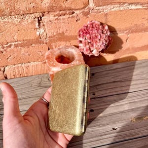 May include: A rectangular, gold-colored metal box with a textured surface. The box is held in a hand, with a brick wall and wooden surface in the background. A pink crystal and a salt lamp are also visible.