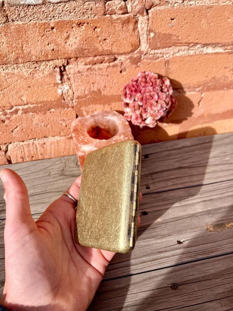 May include: A rectangular, gold-colored metal box with a textured surface. The box is held in a hand, with a brick wall and wooden surface in the background. A pink crystal and a salt lamp are also visible.