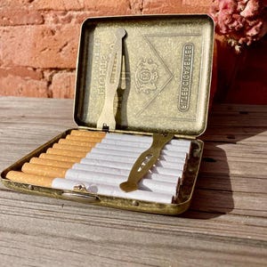 May include: An open, antique-style cigarette case made of brass, filled with cigarettes. The case has a decorative design on the inside lid and a brass cigarette holder. The case is resting on a wooden surface.