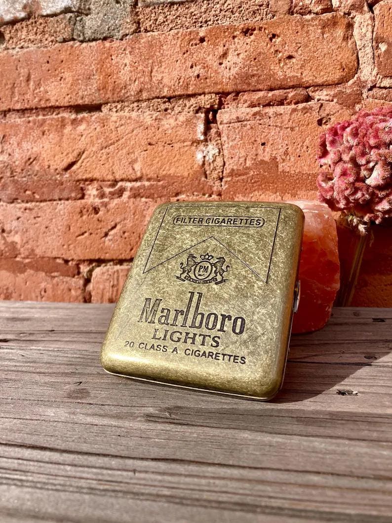 May include: A vintage gold-toned cigarette case with the Marlboro Lights logo and text. The case is rectangular and has a textured surface. The background includes a weathered brick wall and a wooden surface.