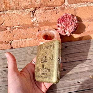 May include: A gold-colored metal cigarette case with the Marlboro Lights logo and text. The case is held in a hand, with a brick wall and decorative items in the background. The case is rectangular and has a hinged lid.