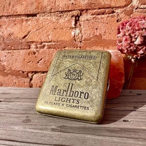 May include: A vintage gold-toned cigarette case with the Marlboro Lights logo and text. The case is rectangular and has a textured surface. The background includes a brick wall, a wooden surface, and a pink flower.