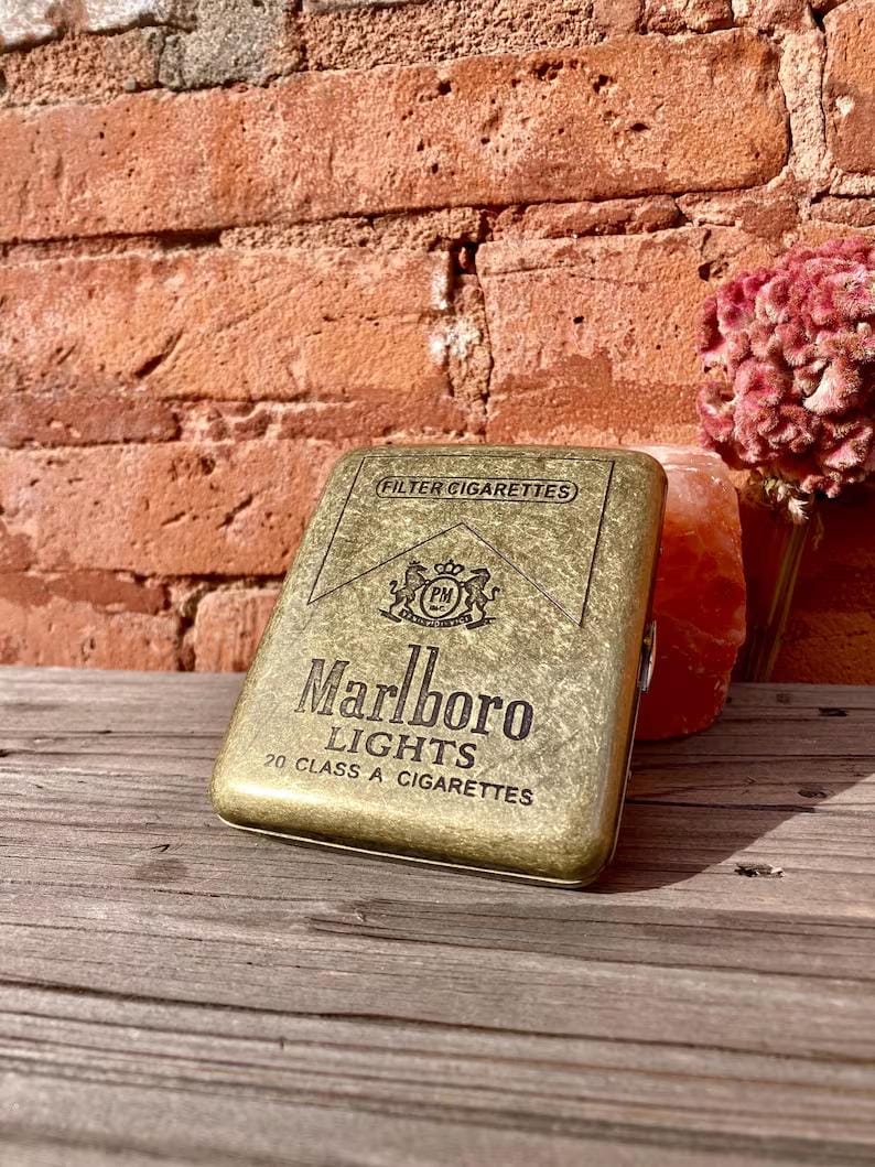 May include: A vintage gold-toned cigarette case with the Marlboro Lights logo and text. The case is rectangular and has a textured surface. The background includes a brick wall, a wooden surface, and a pink flower.
