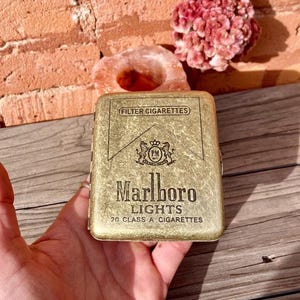 May include: A gold-colored Marlboro Lights cigarette case. The case has the words "FILTER CIGARETTES" at the top, a crest in the middle, and "Marlboro LIGHTS 20 CLASS A CIGARETTES" at the bottom. The case is held in a hand.