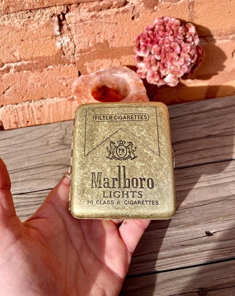 May include: A gold-colored Marlboro Lights cigarette case. The case has the words "FILTER CIGARETTES" at the top, a crest in the middle, and "Marlboro LIGHTS 20 CLASS A CIGARETTES" at the bottom. The case is held in a hand.