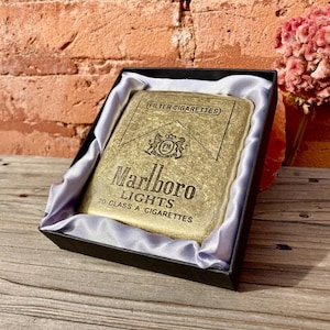 May include: A gold-colored cigarette case with the "Marlboro Lights" logo and text, presented in a black box with a satin lining. The case is rectangular and features the words "FILTER CIGARETTES" at the top.