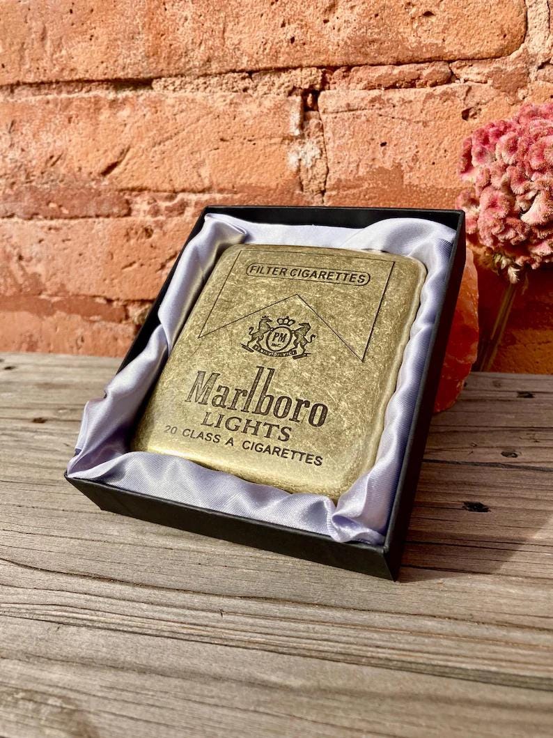 May include: A gold-colored cigarette case with the "Marlboro Lights" logo and text, presented in a black box with a satin lining. The case is rectangular and features the words "FILTER CIGARETTES" at the top.