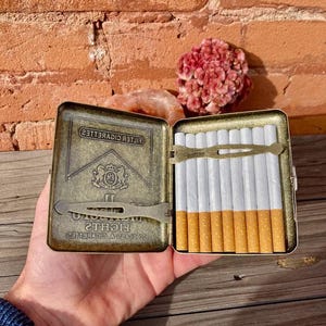 May include: An open, antique-style cigarette case with a brass finish, filled with cigarettes. The case has the words "FILTER CIGARETTES" and "LIGHTS" printed on it. The case is held open in a person's hand.