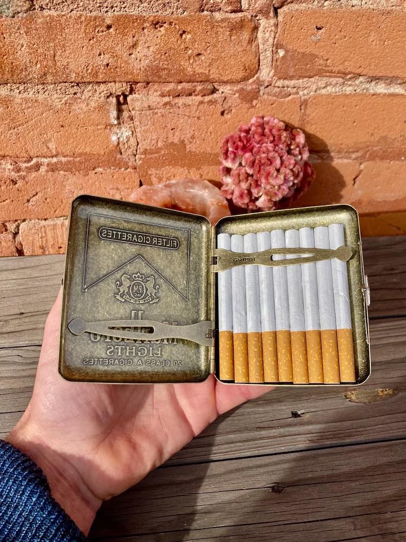 May include: An open, antique-style cigarette case with a brass finish, filled with cigarettes. The case has the words "FILTER CIGARETTES" and "LIGHTS" printed on it. The case is held open in a person's hand.