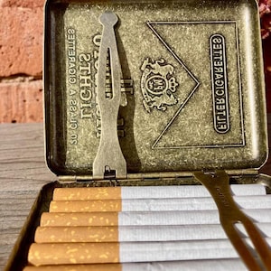 May include: An open, antique-style cigarette case with a brass finish, filled with cigarettes. The case has a hinged lid with embossed text and a decorative emblem. A metal tool rests inside the case. The cigarettes have white filters and brown tobacco.