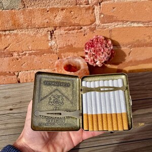 May include: An open, vintage cigarette case with a brass finish, holding a row of cigarettes. The case has the brand name "Lights" and a logo. The cigarettes have white paper and brown filters.