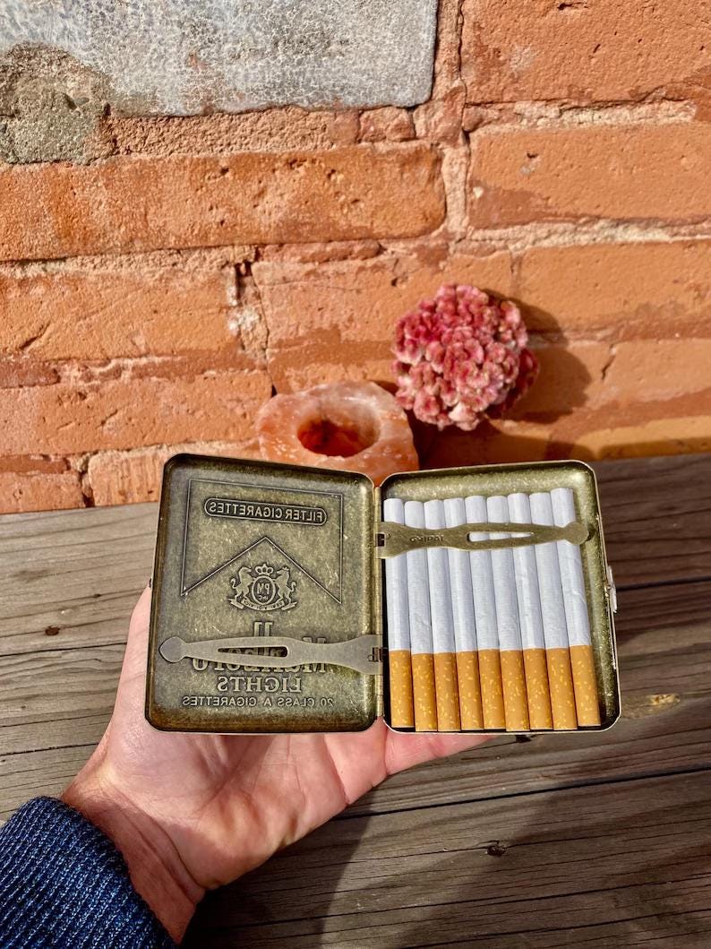 May include: An open, vintage cigarette case with a brass finish, holding a row of cigarettes. The case has the brand name "Lights" and a logo. The cigarettes have white paper and brown filters.