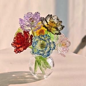 Könnte beinhalten: Eine kleine Glasvase enthält einen Strauß bunter Glasblumen. Die Blumen sind in den Farben Rot, Lila, Gelb, Blau, Grün und Rosa gehalten. Die Vase ist klar und rund, mit grünen Glasstielen. Die Blumen sind detailliert und filigran.