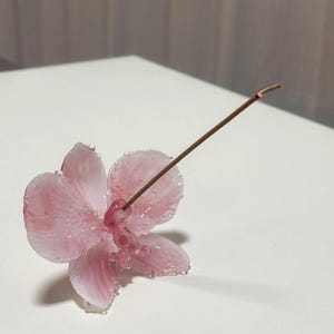 May include: A pink orchid-shaped incense holder with a lit incense stick. The flower has delicate petals with water droplets, and the incense stick is brown. The holder is on a white surface.
