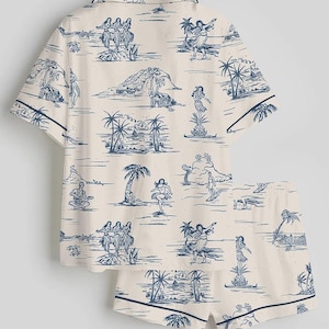 May include: A two-piece pajama set featuring a cream-colored background with a blue island-themed print. The set includes a short-sleeved button-up shirt and matching shorts, both trimmed with a dark blue border. The print depicts palm trees, dancers, and musicians.