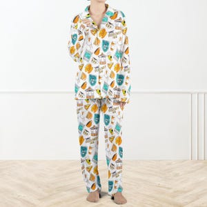Op de afbeelding: Witte pyjamaset met een herhalend patroon van cartoon illustraties in oranje, blauwgroen en geel. De illustraties omvatten koffiebekers, stoelen en de tekst "Dragonfly Inn". De pyjama top heeft een kraag en een knoopsluiting.