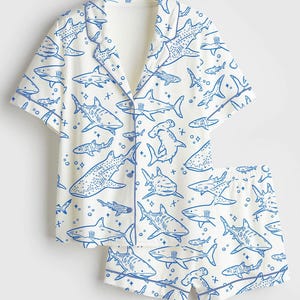 May include: White pajama set with a blue shark pattern. The set includes a short-sleeved button-up shirt and matching shorts. The sharks are in various poses and sizes, creating a playful design. The edges of the shirt and shorts are trimmed in blue.