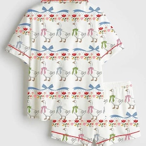 Może przedstawiać: Biały zestaw piżamy z fantazyjnym wzorem gęsi. Kr&oacute;tki rękaw i szorty mają powtarzający się wz&oacute;r gęsi w kolorowych kokardach, przeplatany sercami, kwiatami i niebieskimi wstążkami. Czerwone lam&oacute;wki zdobią krawędzie.