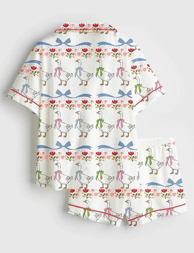 Może przedstawiać: Biały zestaw piżamy z fantazyjnym wzorem gęsi. Kr&oacute;tki rękaw i szorty mają powtarzający się wz&oacute;r gęsi w kolorowych kokardach, przeplatany sercami, kwiatami i niebieskimi wstążkami. Czerwone lam&oacute;wki zdobią krawędzie.