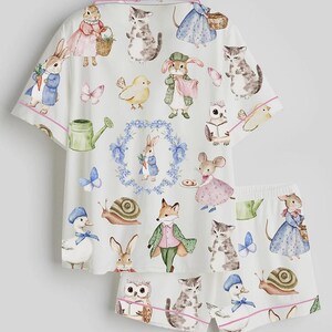 May include: White pajama set featuring a whimsical print of various animals in a storybook style. The set includes a short-sleeved top and matching shorts, both trimmed with pink piping. The print includes rabbits, cats, ducks, and other creatures.