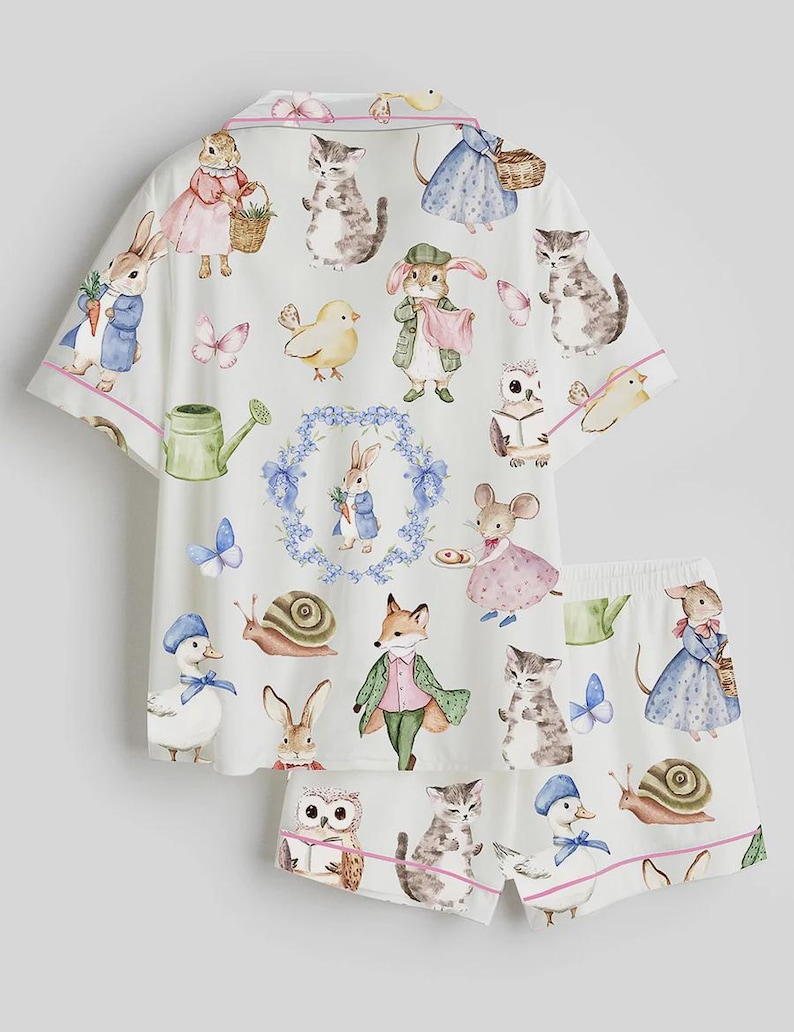 May include: White pajama set featuring a whimsical print of various animals in a storybook style. The set includes a short-sleeved top and matching shorts, both trimmed with pink piping. The print includes rabbits, cats, ducks, and other creatures.