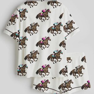 K&ouml;nnte beinhalten: Wei&szlig;es Pyjama-Set mit einem sich wiederholenden Muster von Rennpferden und Jockeys. Das kurz&auml;rmelige Oberteil und die Shorts haben braune Paspeln. Die Jockeys tragen farbige Seiden, und die Pferde sind braun.