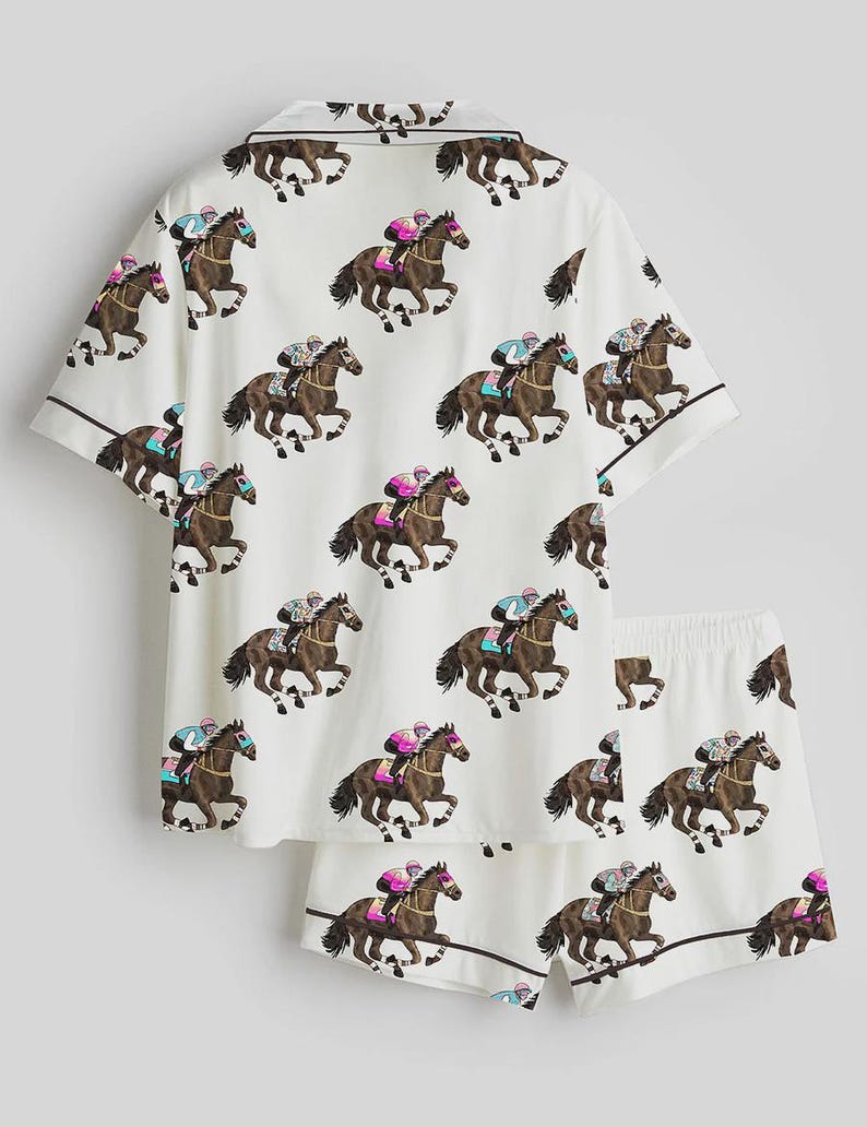 K&ouml;nnte beinhalten: Wei&szlig;es Pyjama-Set mit einem sich wiederholenden Muster von Rennpferden und Jockeys. Das kurz&auml;rmelige Oberteil und die Shorts haben braune Paspeln. Die Jockeys tragen farbige Seiden, und die Pferde sind braun.