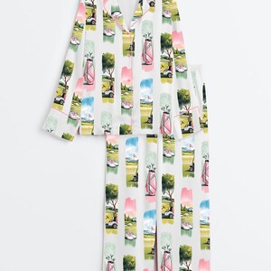 Peut inclure: Ensemble de pyjama blanc avec un motif sur le th&egrave;me du golf. Le motif pr&eacute;sente des rayures verticales avec des illustrations de sacs de golf, de voiturettes de golf, de chaussures de golf et d'arbres en rose, vert, bleu et jaune. L'ensemble comprend un haut boutonn&eacute; et un pantalon assorti.