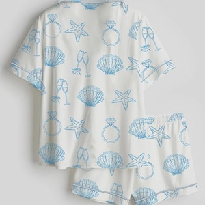 Op de afbeelding: Witte pyjamaset met een blauw patroon van zeesterren, schelpen, champagneglazen en diamanten ringen. De set bestaat uit een overhemd met korte mouwen en bijpassende shorts. De randen zijn afgewerkt met een dunne blauwe rand.
