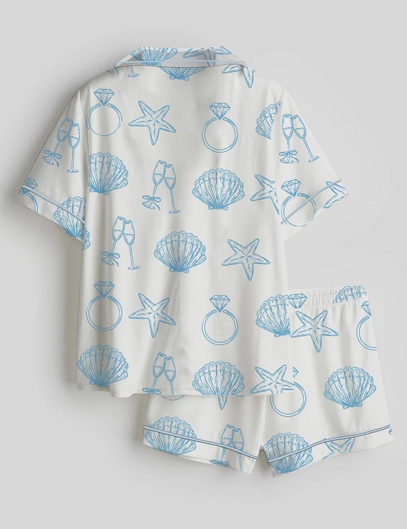 Op de afbeelding: Witte pyjamaset met een blauw patroon van zeesterren, schelpen, champagneglazen en diamanten ringen. De set bestaat uit een overhemd met korte mouwen en bijpassende shorts. De randen zijn afgewerkt met een dunne blauwe rand.