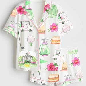 May include: White pajama set with a golf-themed pattern. The set includes a short-sleeved button-up shirt and shorts, both adorned with golf carts, golf balls, sandwiches, pink flowers, and golf bags. The trim is pink.