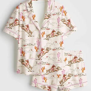 Könnte beinhalten: Cremefarbenes Pyjama-Set mit Cowboy- und Pferdemuster. Das kurzärmelige Oberteil und die Shorts zeigen ein sich wiederholendes Design von Cowboys, Pferden, Kakteen und Wolken. Die Kanten sind mit rosa Paspeln versehen.