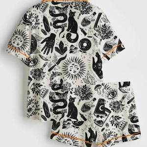 May include: White pajama set with a black occult-themed print featuring suns, snakes, cats, crystals, and hands. The short-sleeved top and shorts have orange trim.