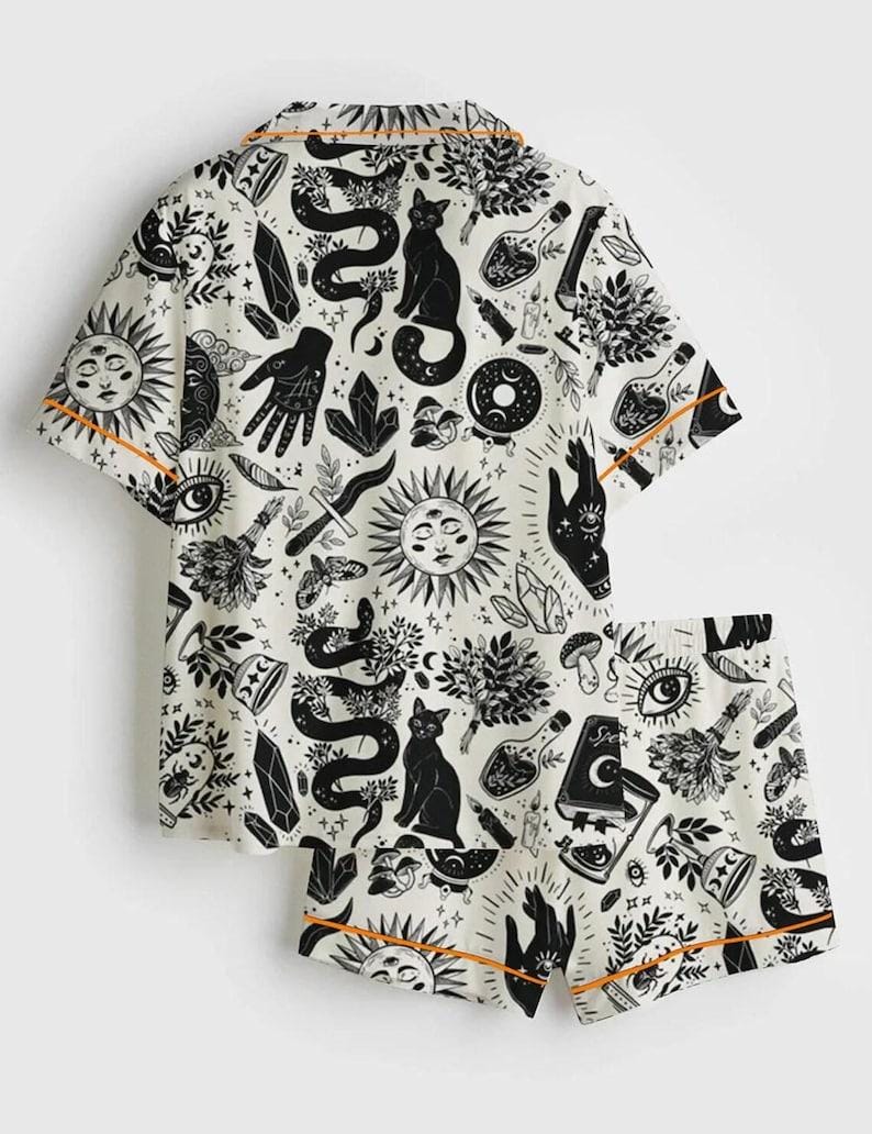 May include: White pajama set with a black occult-themed print featuring suns, snakes, cats, crystals, and hands. The short-sleeved top and shorts have orange trim.