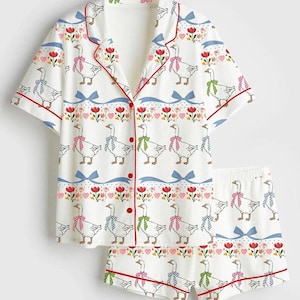 Może przedstawiać: Biały zestaw piżamowy z fantazyjnym nadrukiem gęsi. Kr&oacute;tki rękaw topu i szorty mają powtarzający się wz&oacute;r gęsi w kokardkach, czerwone wykończenia i kwiatowe akcenty. Zestaw jest zapinany na guziki.