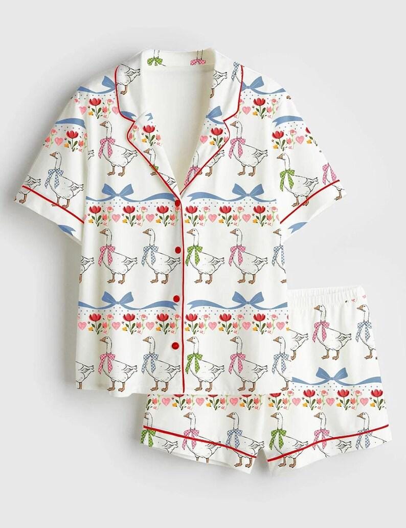 Może przedstawiać: Biały zestaw piżamowy z fantazyjnym nadrukiem gęsi. Kr&oacute;tki rękaw topu i szorty mają powtarzający się wz&oacute;r gęsi w kokardkach, czerwone wykończenia i kwiatowe akcenty. Zestaw jest zapinany na guziki.