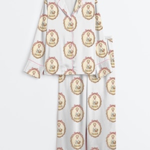 Może przedstawiać: Biały zestaw piżamowy z powtarzającym się wzorem dziewczynki trzymającej kosz w owalnej ramie. Zestaw zawiera koszulę zapinaną na guziki i spodnie, wykończone r&oacute;żową lam&oacute;wką. Piżama wykonana jest z miękkiego, jedwabistego materiału.