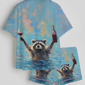 Puede incluir: Conjunto de pijama azul claro con un mapache sosteniendo una copa de vino y una botella de vino en el agua. La camisa y el pantal&oacute;n corto tienen ribetes rosas. El mapache es el foco central del dise&ntilde;o.