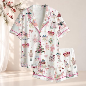 May include: A white short-sleeve button-down pajama shirt and matching shorts set featuring a colorful holiday-themed print of nutcrackers, ballerinas, Christmas trees, crowns, castles, and gift boxes with pink piping along the edges.