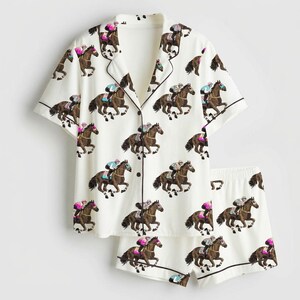 K&ouml;nnte beinhalten: Wei&szlig;es Pyjama-Set mit einem sich wiederholenden Muster aus braunen Pferden und Jockeys in verschiedenen Farben. Das kurz&auml;rmelige Oberteil und die Shorts haben schwarze Paspeln. Das Pyjama-Set ist aus einem weichen, leichten Stoff gefertigt.
