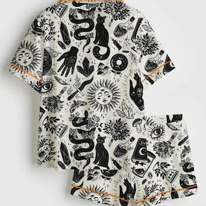 May include: A two-piece pajama set featuring a white background with a black occult-themed pattern. The pattern includes suns, snakes, cats, hands, crystals, and potion bottles. The set has orange trim.