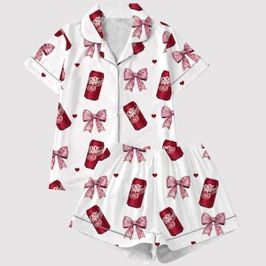 May include: White pajama set with a pattern of red Dr Pepper cans, pink bows, and red hearts. The set includes a short-sleeved button-up shirt and matching shorts. The pajama set is made of a soft, comfortable material.