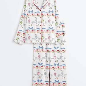 Może przedstawiać: Biały zestaw piżamy z powtarzającym się wzorem gęsi w kokardkach, niebieskich kokardach i kwiatowych akcentach. Top z długim rękawem ma czerwone wykończenia i guziki. Spodnie mają pasujący wz&oacute;r i czerwone wykończenia.