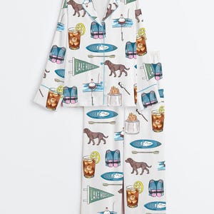 Peut inclure: Ensemble de pyjama blanc avec un motif r&eacute;p&eacute;t&eacute; d'illustrations sur le th&egrave;me du lac. Le motif comprend des chiens bruns, des gilets de sauvetage, des kayaks, des flotteurs de p&ecirc;che, des cocktails au citron et des fanions avec l'inscription "LAKE HOUSE".
