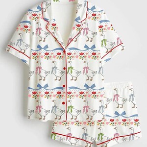 Może przedstawiać: Biały zestaw piżamowy z fantazyjnym nadrukiem gęsi. Kr&oacute;tki rękaw topu i szorty mają powtarzający się wz&oacute;r gęsi w kokardkach, czerwonych wykończeniach i kwiatowych akcentach. Zestaw zawiera koszulę zapinaną na guziki i szorty.