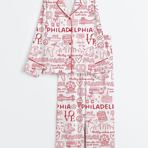 May include: White pajama set with a red Philadelphia-themed print. The set includes a button-down top and pants, featuring city landmarks, icons, and text like "Philadelphia" and "Pennsylvania". The pajamas have red trim.
