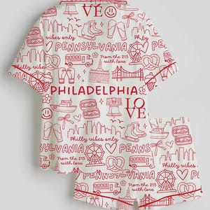 May include: White pajama set with a red Philadelphia-themed print. The set includes a short-sleeved top and shorts, both featuring city landmarks, icons, and text. The trim is red.
