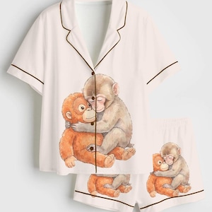May include: White pajama set with a short-sleeved top and shorts. The set features a brown trim and a print of two monkeys hugging. The top has a button-down front and a notched collar. The shorts have an elastic waistband.
