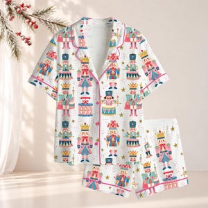 May include: A white short-sleeve button-down pajama shirt and matching shorts set featuring a repeating pattern of colorful nutcracker figures, drums, and gold stars. The garment has pink piping along the collar, front placket, and hem.