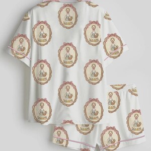 Może przedstawiać: Biały zestaw piżamowy z powtarzającym się wzorem ilustracji gęsi w owalnej ramie, z r&oacute;żowymi kokardkami i wykończeniami. Zestaw zawiera top z kr&oacute;tkim rękawem i pasujące szorty.